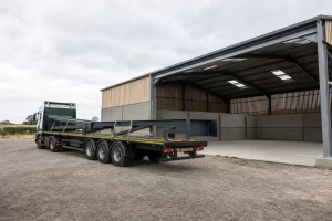 Specialist flatbed carrying a long steel frame section beside a rural steel shed